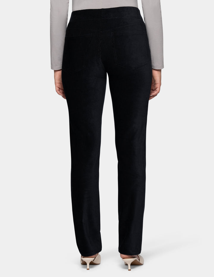 A backside view of a model wearing black corduroy pants and a long sleeve grey top with ivory pointed toe shoes