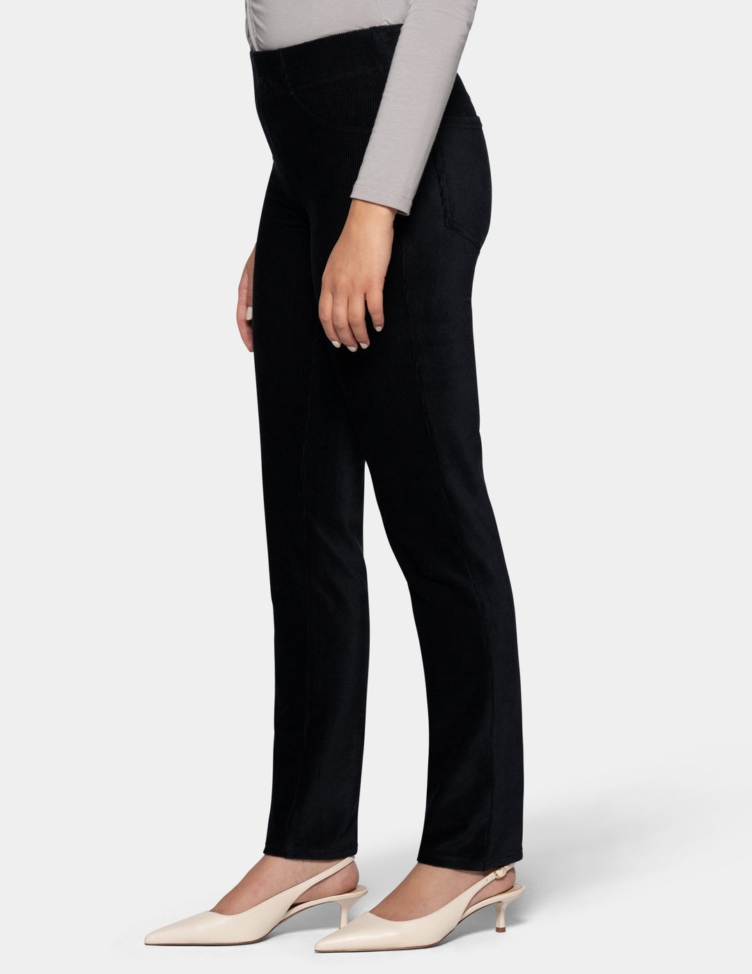 A side view of a model wearing black corduroy pants and a long sleeve grey top with ivory pointed toe shoes