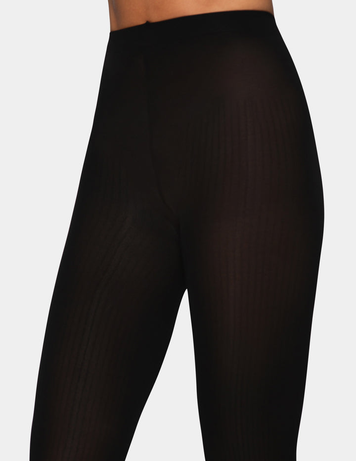 Black ribbed tights shown on a model from the waist down, highlighting the vertical ribbed texture against a light gray background.