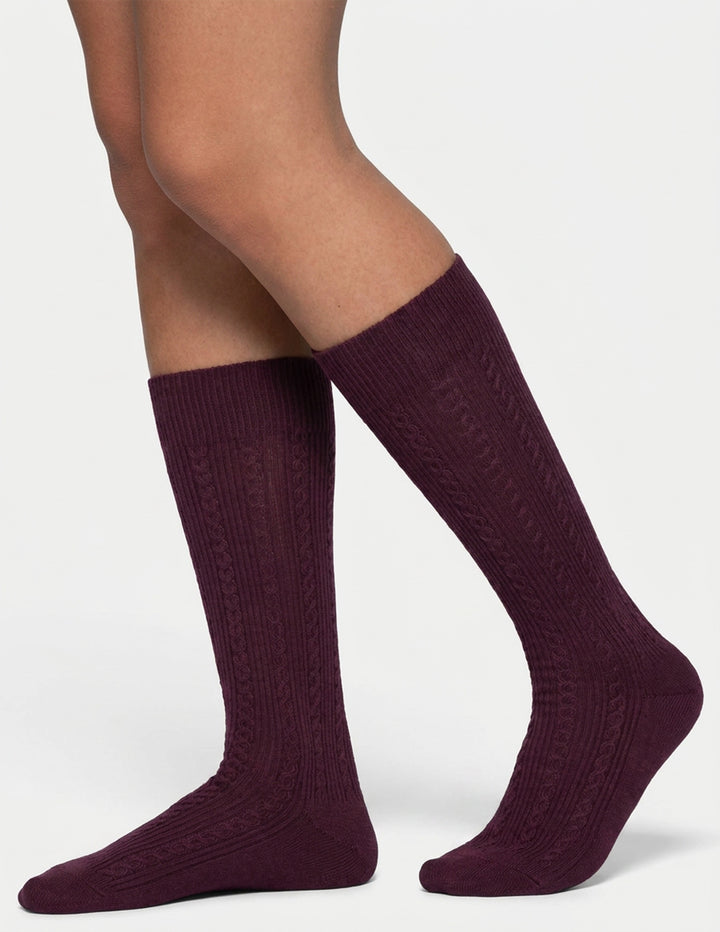 A woman modeling burgundy ribbed knee high socks against a light gray background.