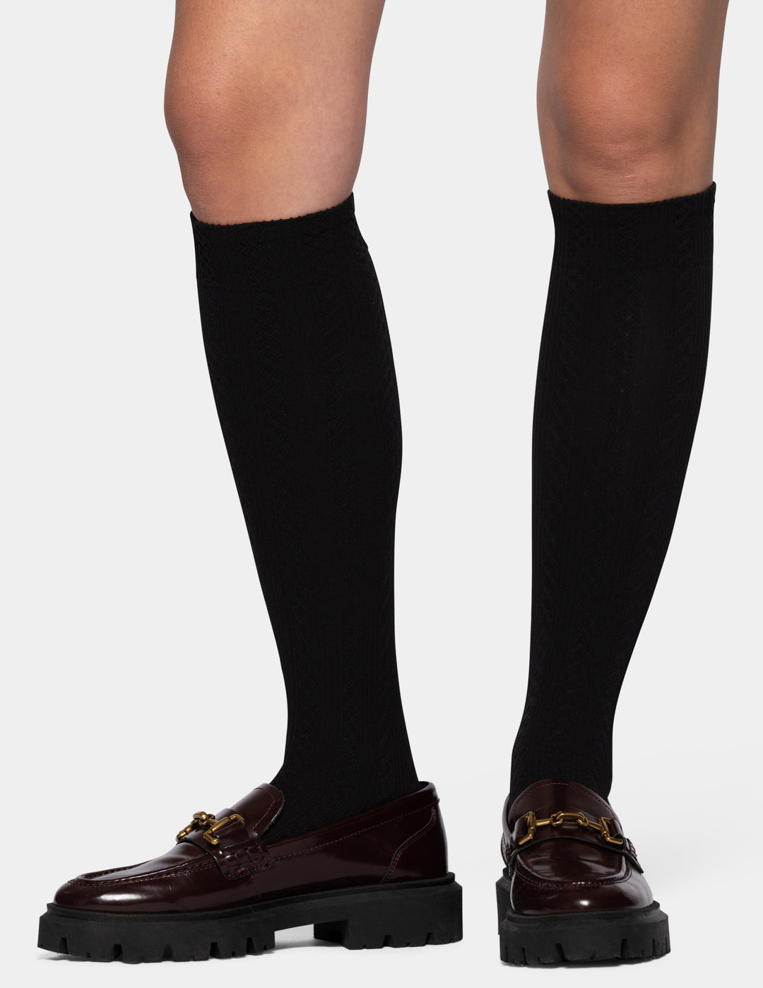A woman wearing black knee-high socks and dark brown patent leather loafers with chunky black soles and gold-tone hardware, shown against a light gray background.