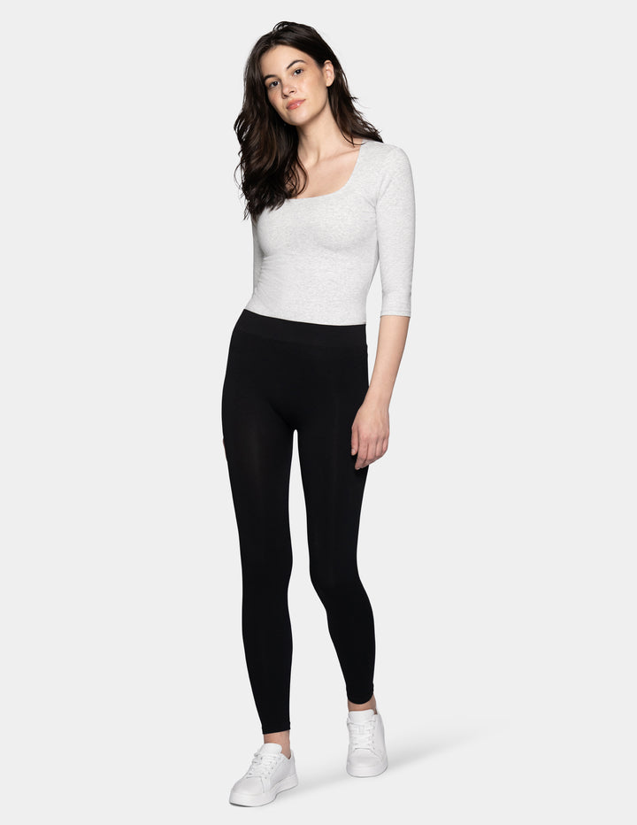 A model wearing black leggings, light grey 3/4 sleeve shirt, and white sneakers photographed against a light gray background.