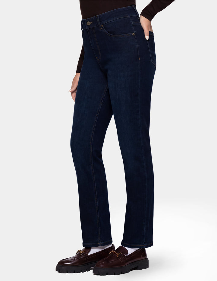 A model wearing dark blue denim pants, a long sleeve brown top, and brown loafers photographed against a light gray background.