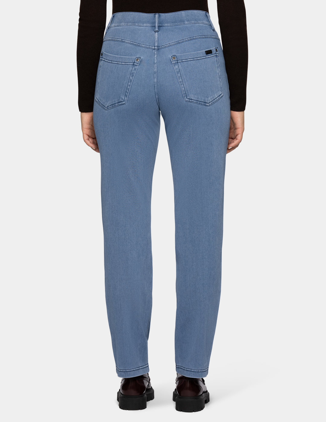 A back view of a model wearing light blue denim pants, a long sleeve black top, and loafers photographed against a light gray background.