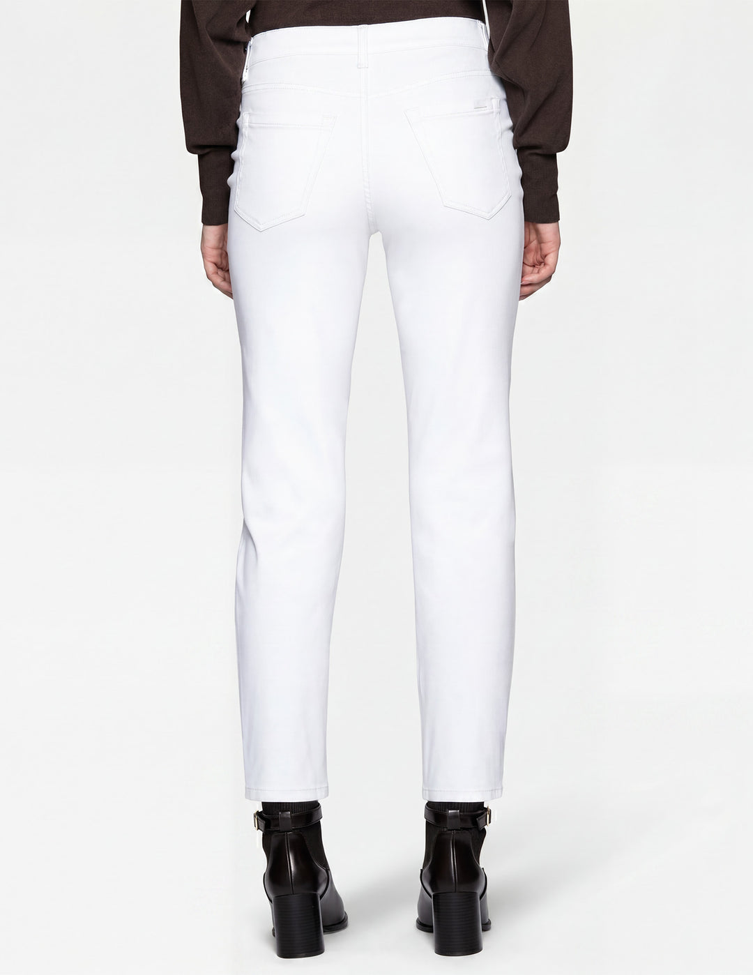 A backside view of a model wearing white pants, a brown long sleeve top, and black boots photographed against a light gray background.