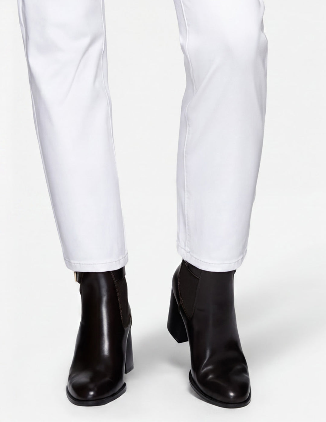 A closeup of a model wearing white pants and black boots photographed against a light gray background.