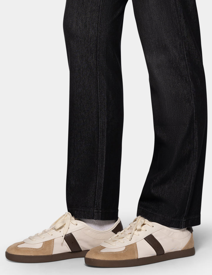 A detailed view of a model wearing black denim pants, a long sleeve white top, and sneakers photographed against a light gray background.