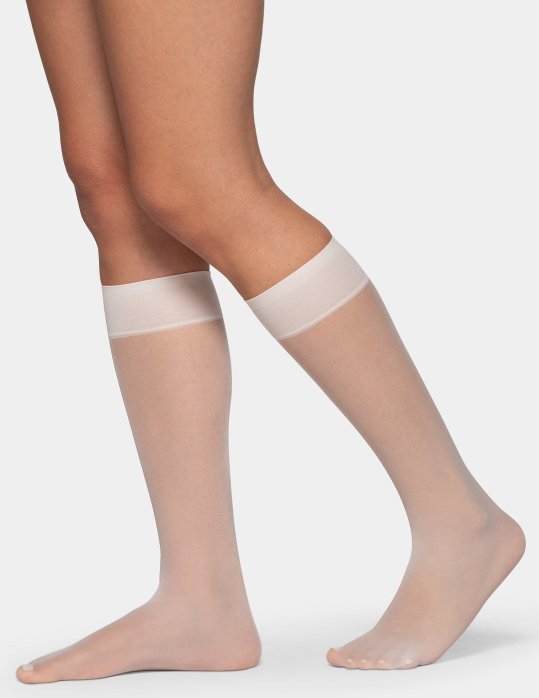 A close-up of a person's legs wearing sheer knee-high stockings with opaque white bands at the top, shown against a light gray studio background.