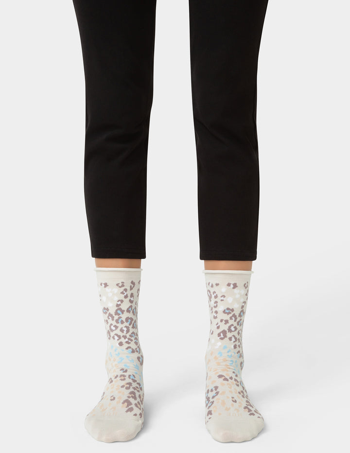 A person's lower legs wearing black cropped leggings and patterned crew socks with a leopard print design in beige, brown, and light blue on a white base, shown against a light gray studio background.