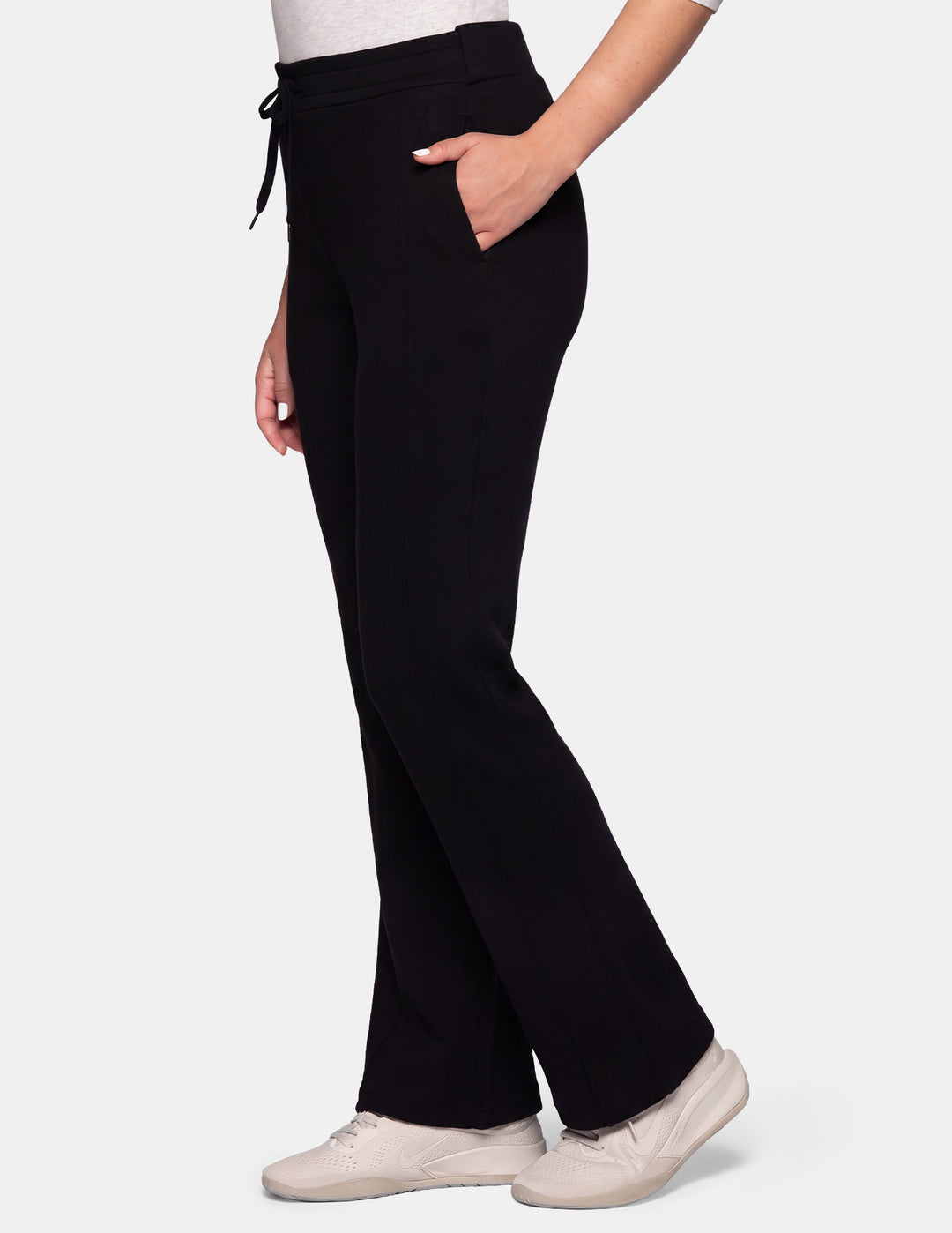 A person wearing black trackpants with side pockets and a drawstring waist, paired with off-white sneakers, shown against a plain light grey studio background.