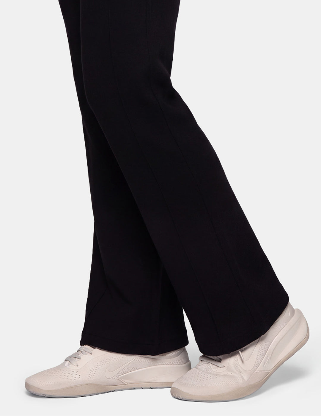 A close-up of a model wearing black jersey trackpants and off-white sneakers against a gray background.