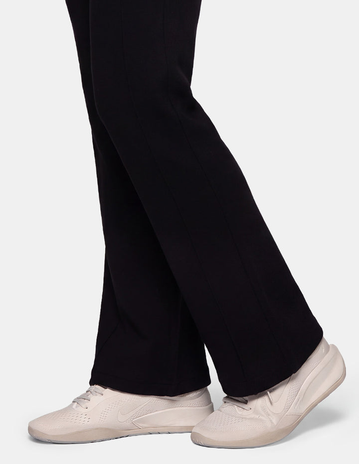 A close-up of a model wearing black jersey trackpants and off-white sneakers against a gray background.