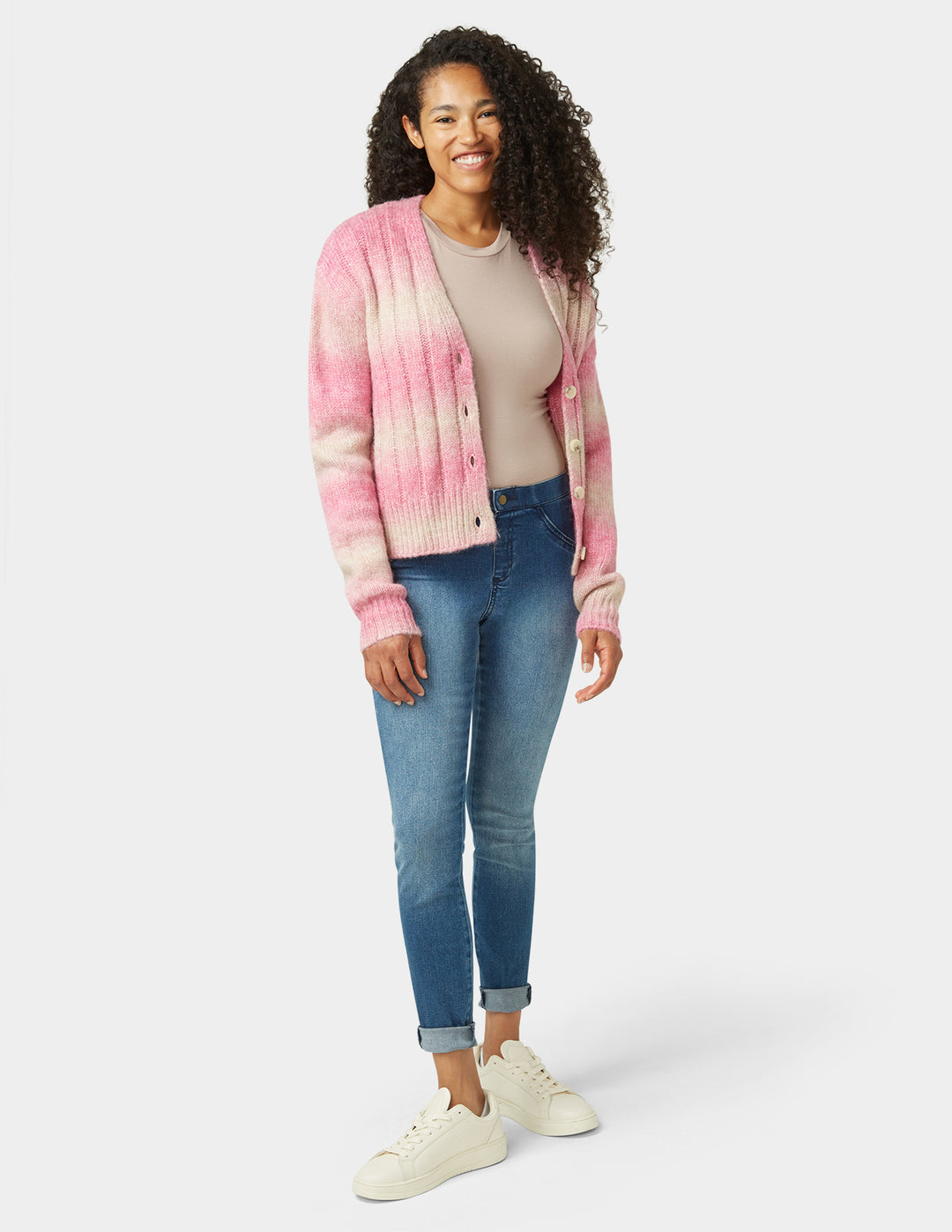 Woman wearing a pink and white striped cardigan, beige top, blue jeans, and white sneakers on a white background