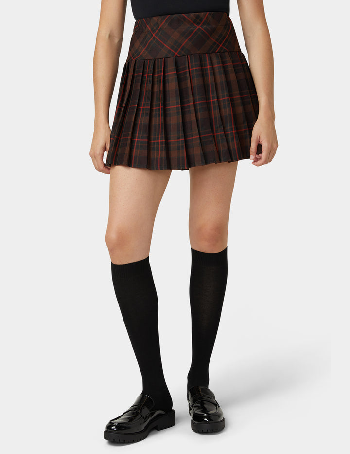 A model wearing a pleated mini skirt in a dark red and black tartan plaid pattern, paired with black knee-high socks and black patent leather loafers, against a light gray studio background.