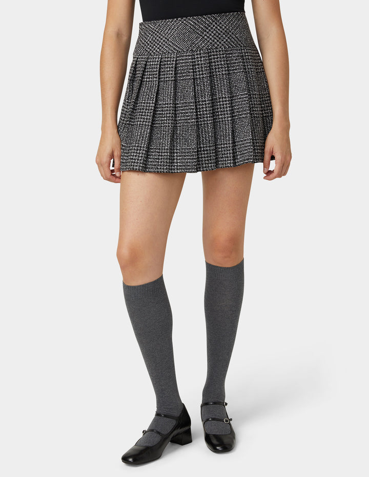 A model wearing a black and white houndstooth pleated mini skirt, dark gray knee-high socks, and black Mary Jane shoes, shown from the waist down on a light gray studio background.