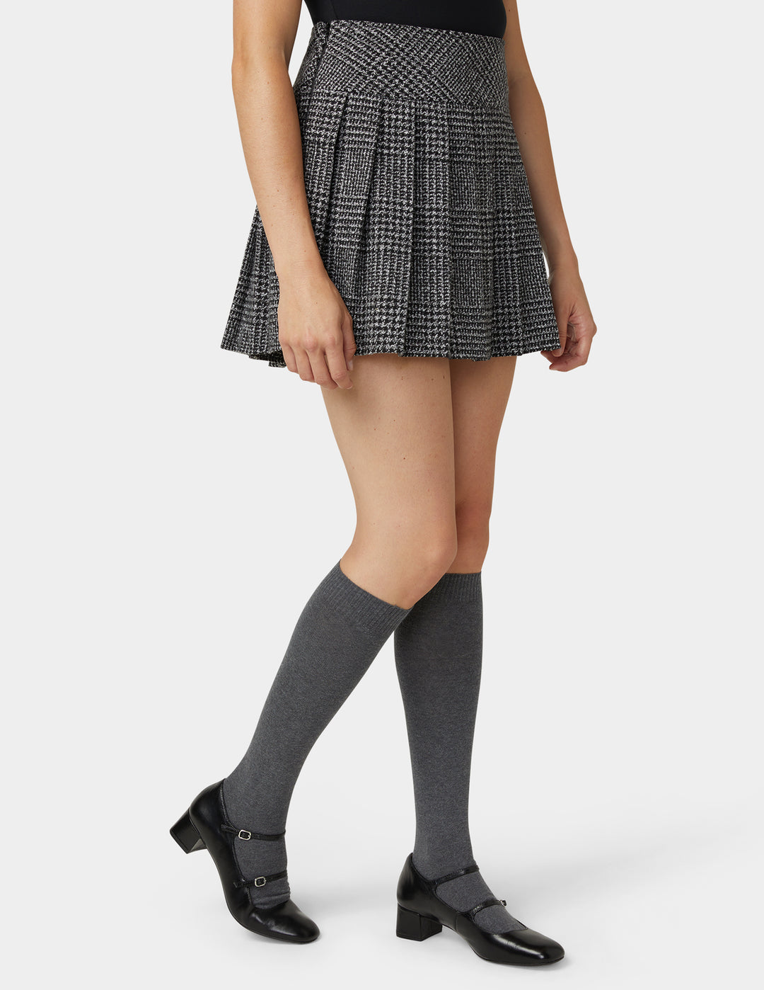 A model wearing a black and white checkered pleated mini skirt, gray knee-high socks, and black Mary Jane heels, shown from the waist down on a light gray studio background.