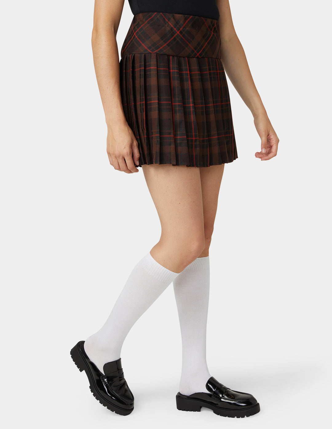 A model wearing a pleated mini skirt in a dark red and black plaid pattern, paired with white knee-high socks and black loafers, against a light gray studio background.