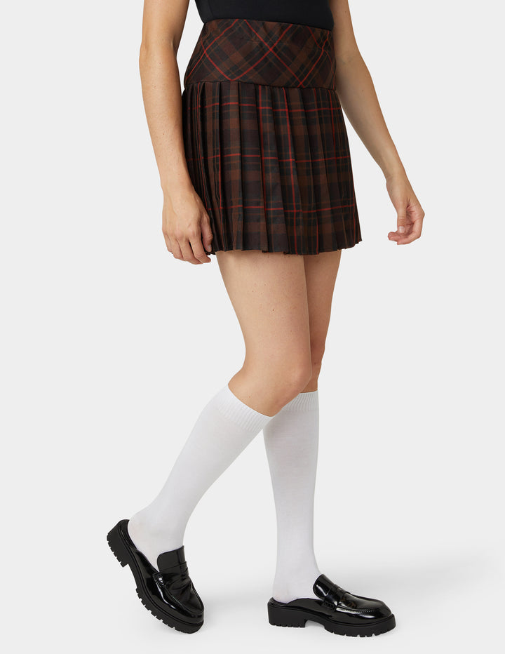 A model wearing a pleated mini skirt in a dark red and black plaid pattern, paired with white knee-high socks and black loafers, against a light gray studio background.