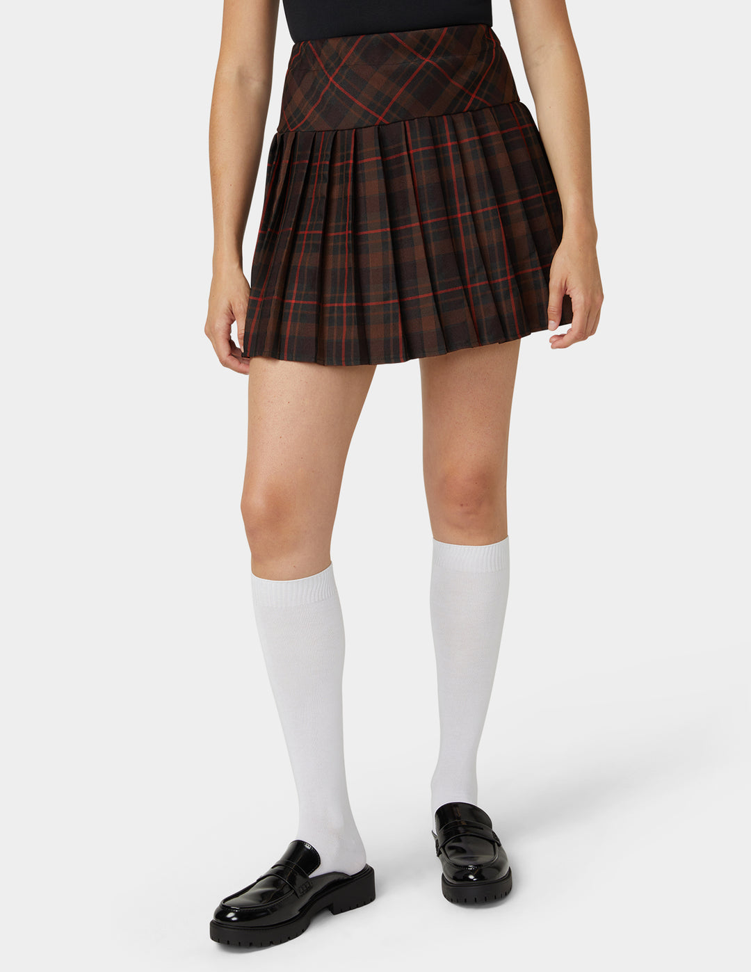 A model wearing a pleated plaid mini skirt in black and red, paired with white knee-high socks and black loafers, shown from the waist down on a light gray background.