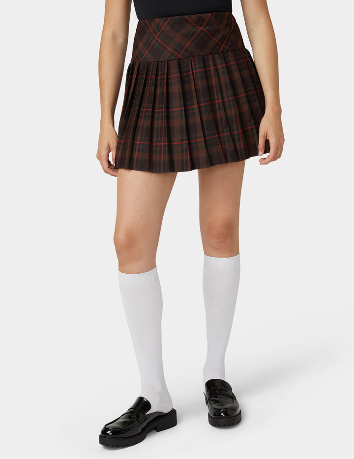 A model wearing a pleated plaid mini skirt in black and red, paired with white knee-high socks and black loafers, shown from the waist down on a light gray background.