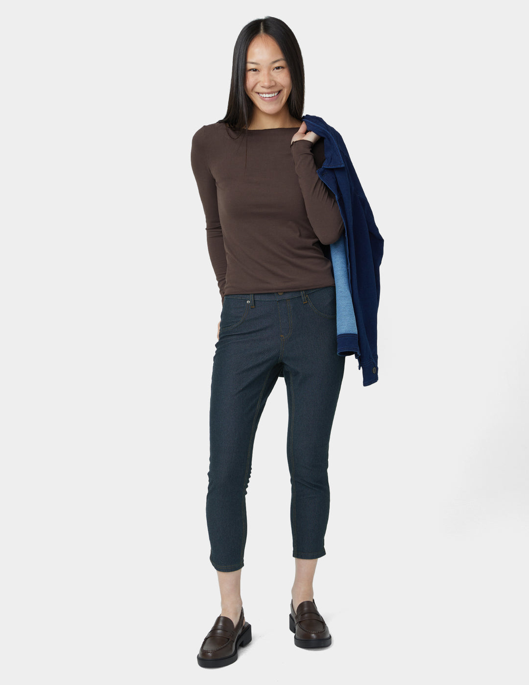 Woman wearing a brown top and dark pants with a blue jacket over her shoulder on a white background