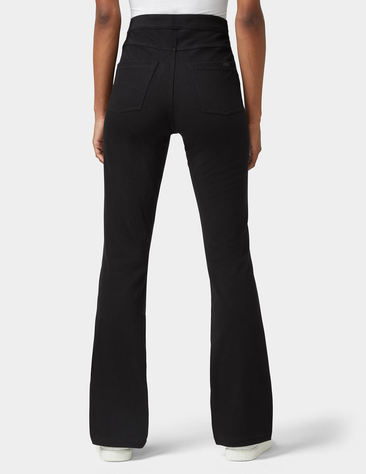 Rear view of a person wearing black flared pants with a white top and white shoes, photographed against a light gray studio background.
