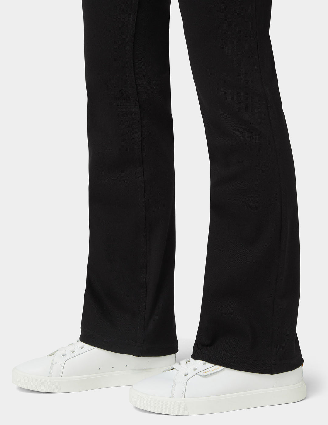 Black flared pants worn with white low-top sneakers, shown from the knees down on a light gray studio background.