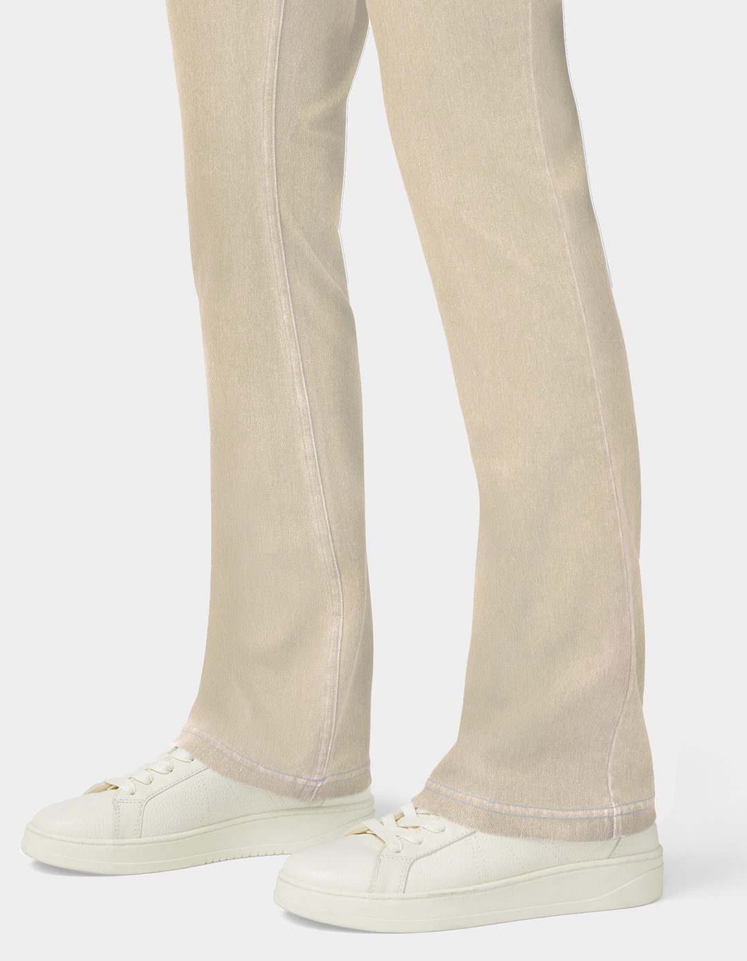 Close-up of a person's legs wearing light beige flared jeans and off-white low-top sneakers against a clean light gray studio background.