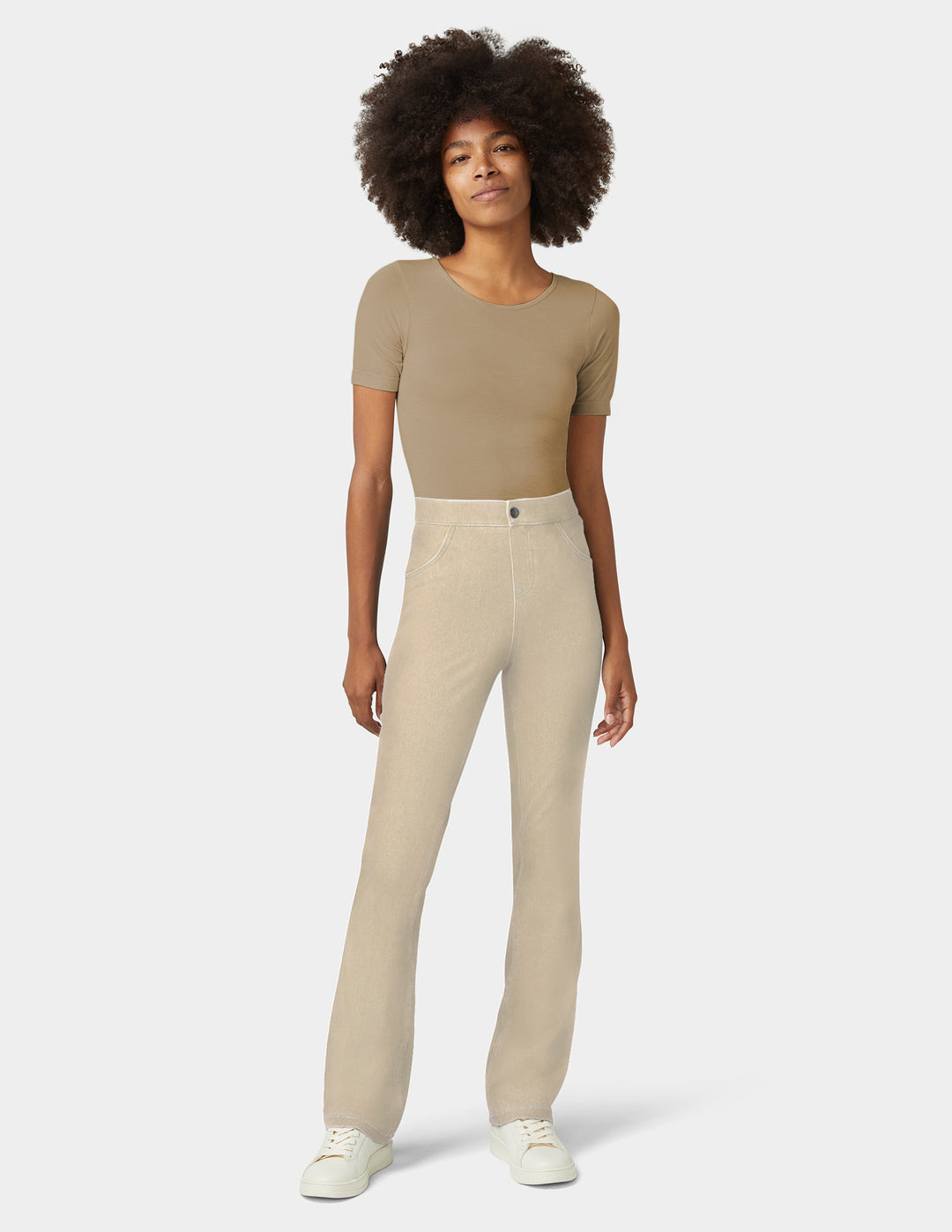 A model against a light gray studio background wearing a beige short-sleeve crop top, light beige straight-leg jeans, and white sneakers.