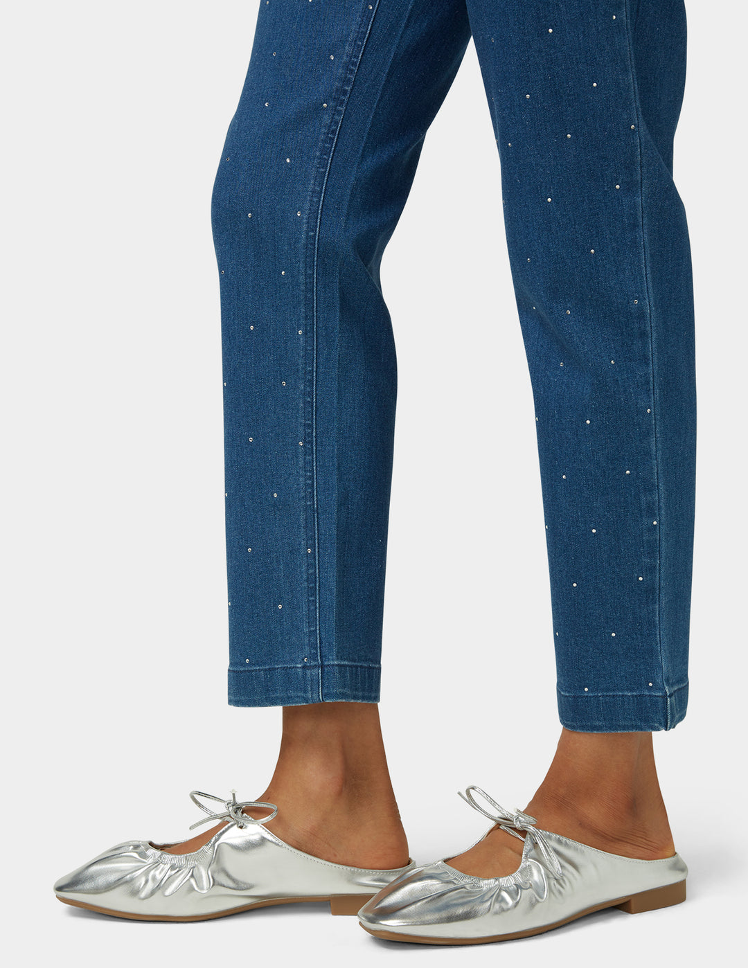 Blue jeans with a subtle pattern worn by a person, paired with silver flats on a white background