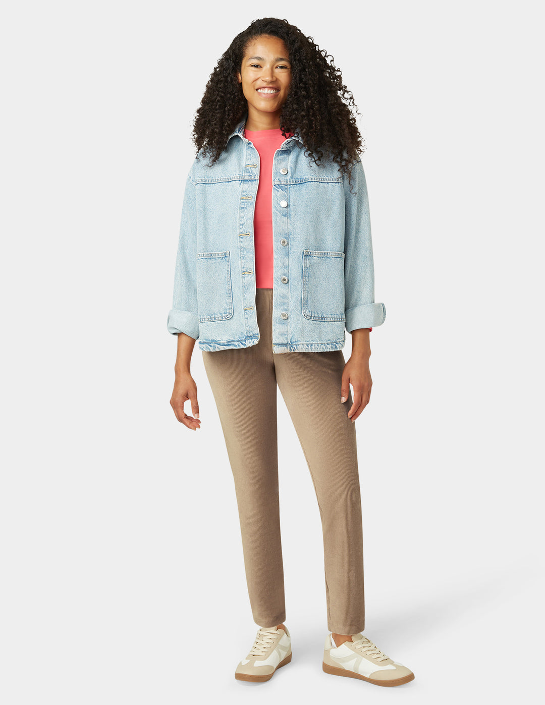Person wearing a light blue denim jacket, pink shirt, beige pants, and white sneakers on a white background