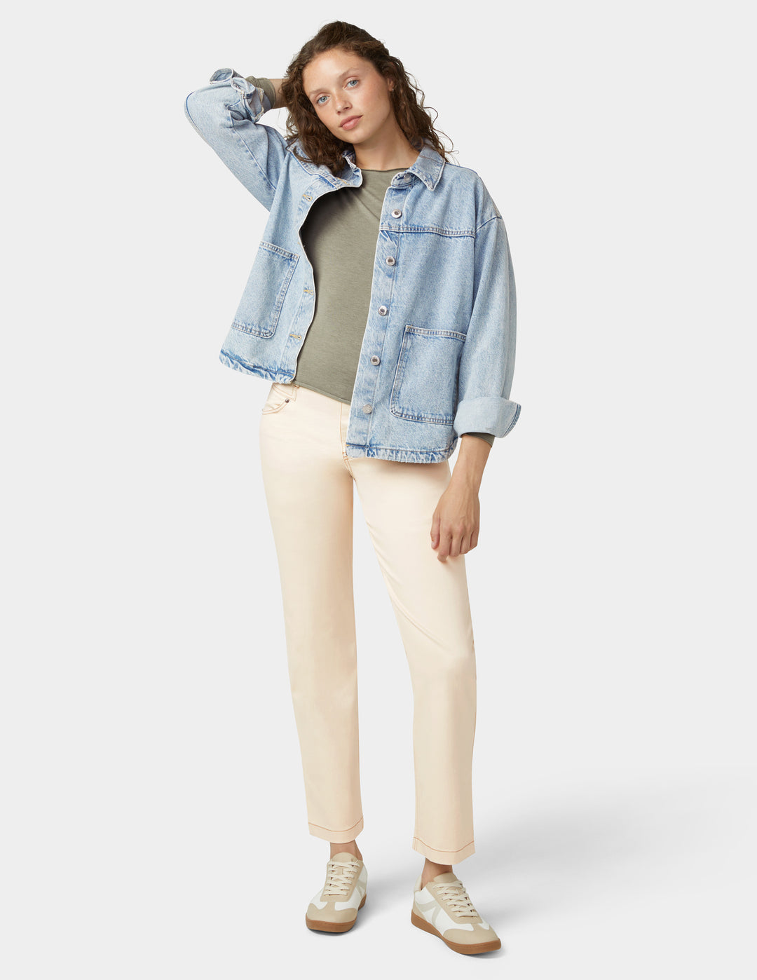 Woman wearing a light blue denim jacket, green shirt, beige pants, and white sneakers on a white background