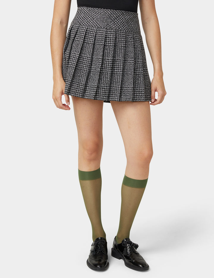Person wearing a plaid skirt, black top, olive green sheer knee-high socks, and black shoes on a white background