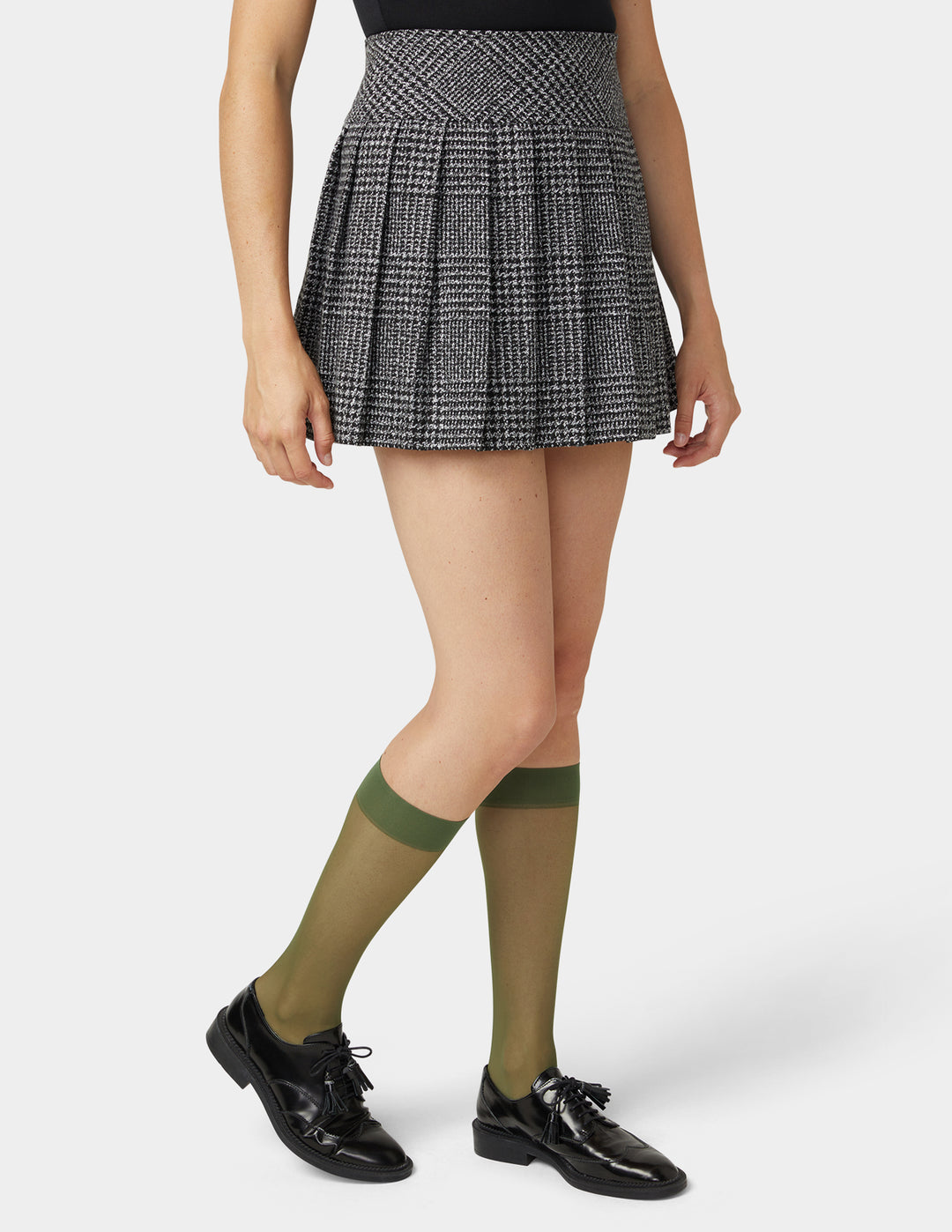 Person wearing a plaid skirt, olive green  sheer knee-high socks, and black shoes on a white background