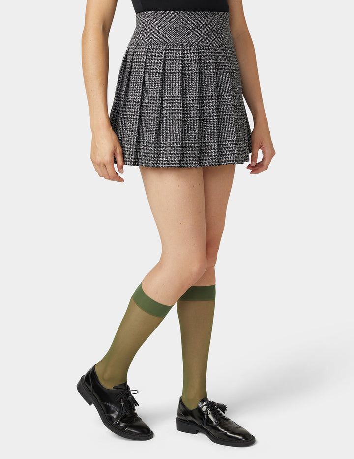 Person wearing a plaid skirt, olive green  sheer knee-high socks, and black shoes on a white background
