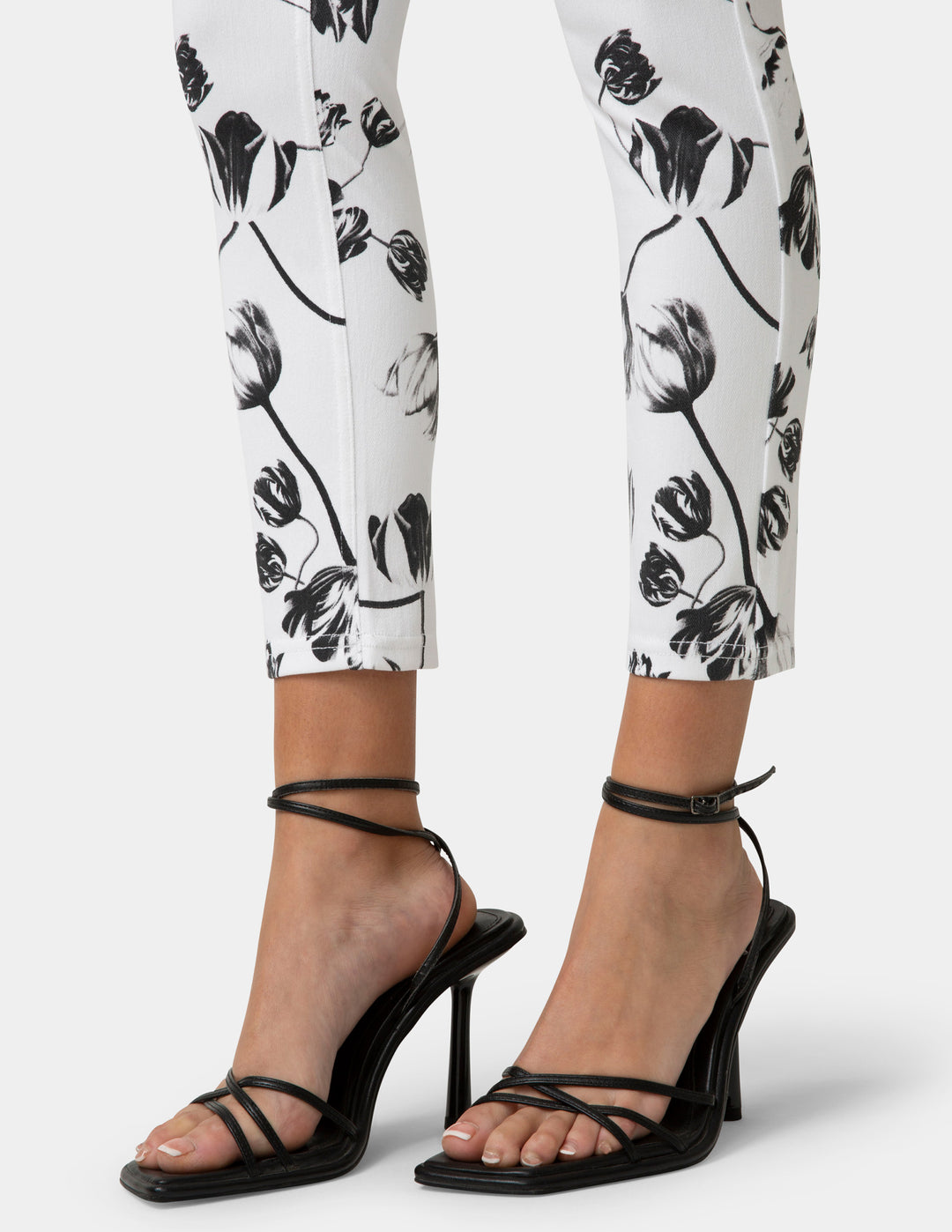 Black strappy high-heeled shoes worn with white pants featuring black floral patterns.